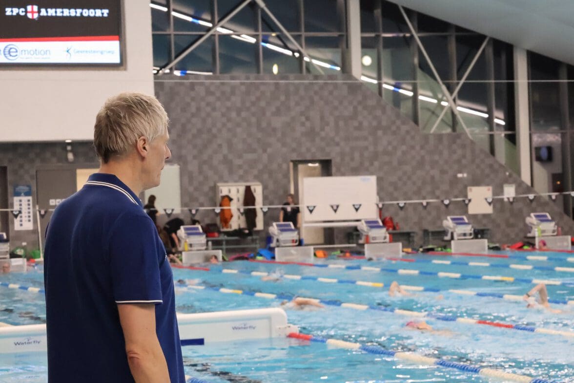 Docent staat aan de badrand van het 50 metyer bad terwijl in het water in meerdere banen de borstcrawl wordt geoefend tijdens de borstcrawlcursus